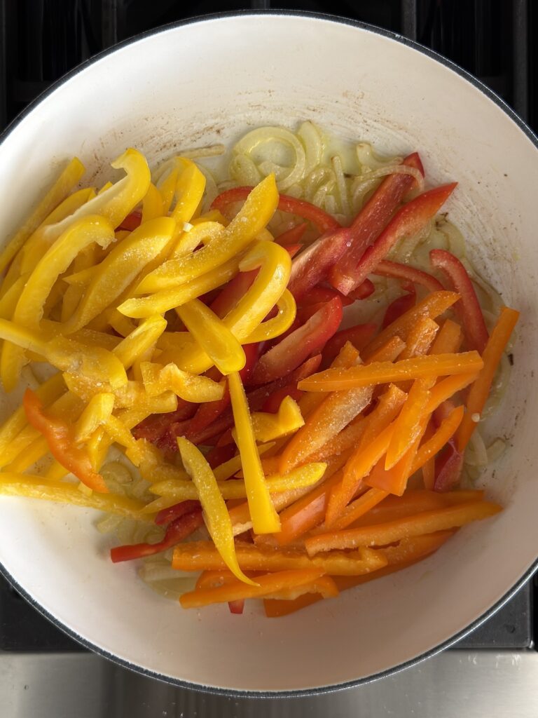 Sliced red, yellow, and orange bell peppers with onions in a white pan – cooking vegetables for Italian pepperonata recipe