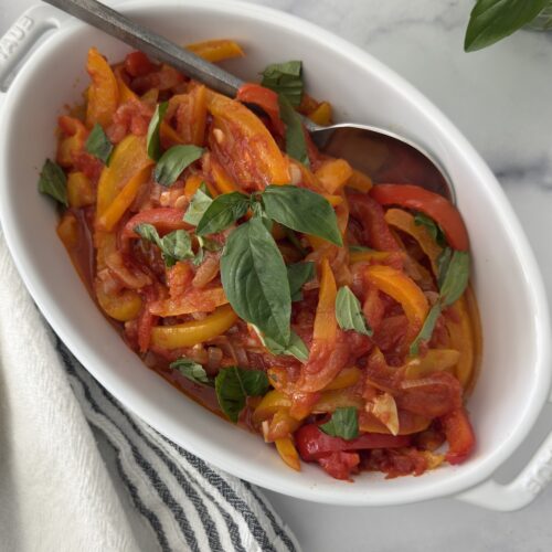 Italian pepperonata served in a white oval dish with fresh basil – sweet peppers and onions simmered with tomato