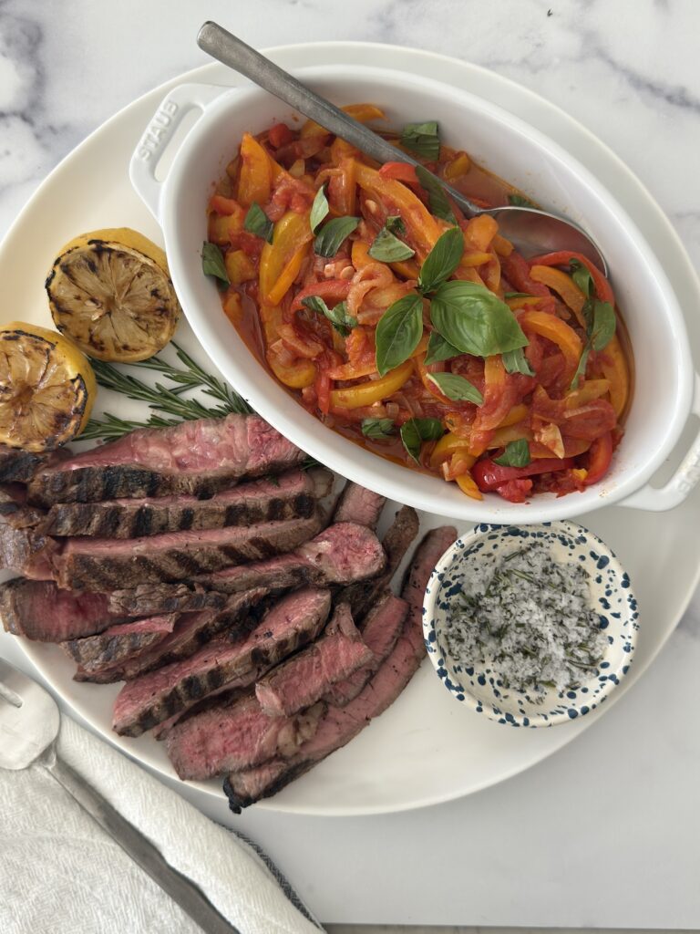 Italian pepperonata served with grilled steak, charred lemon, and fresh herbs – a summer meal idea with sweet peppers and onions