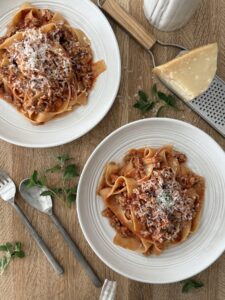 Two bowls of Easy Chicken Bolognese with Italian Sausage served over pappardelle pasta and topped with grated Parmesan, styled on a wooden table with fresh herbs