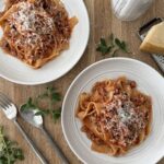 Two bowls of Easy Chicken Bolognese with Italian Sausage served over pappardelle pasta and topped with grated Parmesan, styled on a wooden table with fresh herbs