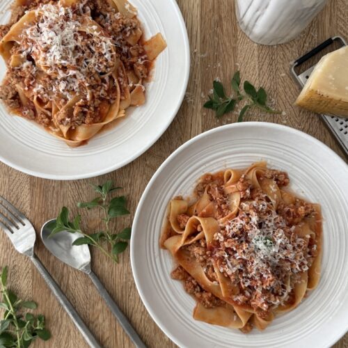 Two bowls of Easy Chicken Bolognese with Italian Sausage served over pappardelle pasta and topped with grated Parmesan, styled on a wooden table with fresh herbs