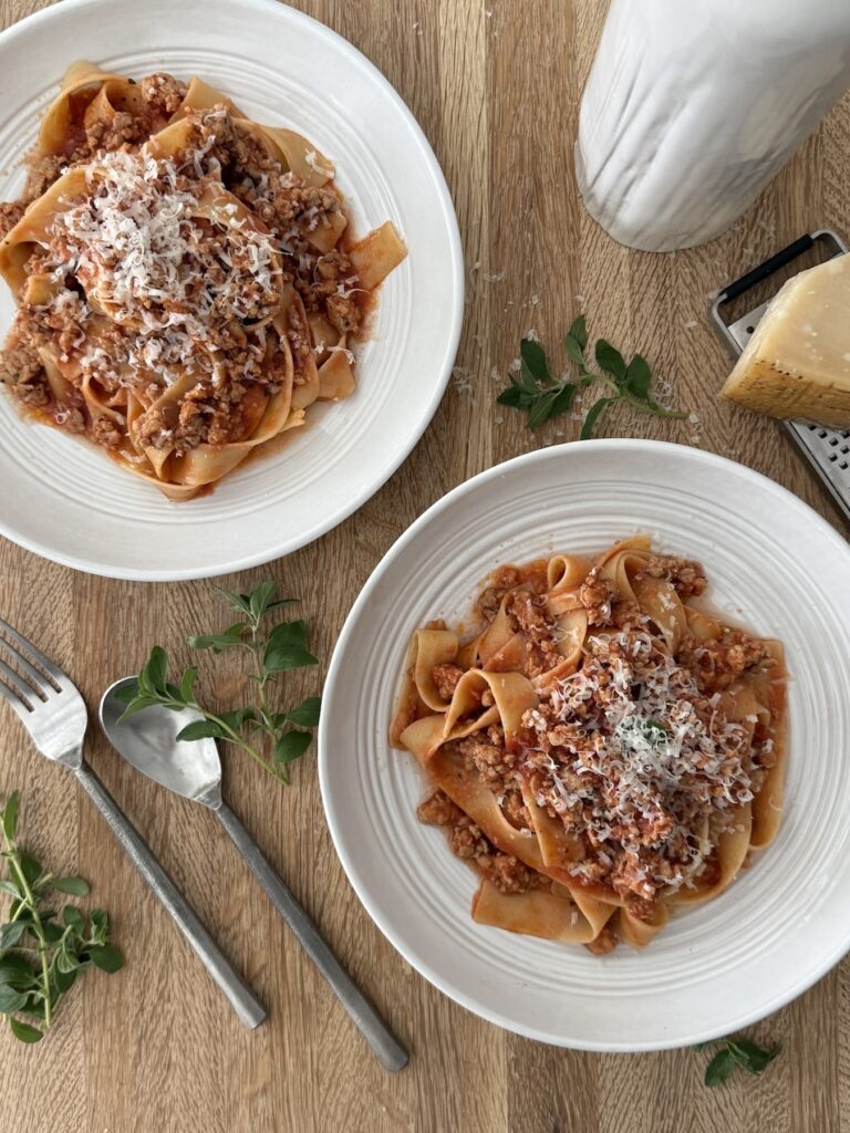 Two bowls of Easy Chicken Bolognese with Italian Sausage served over pappardelle pasta and topped with grated Parmesan, styled on a wooden table with fresh herbs