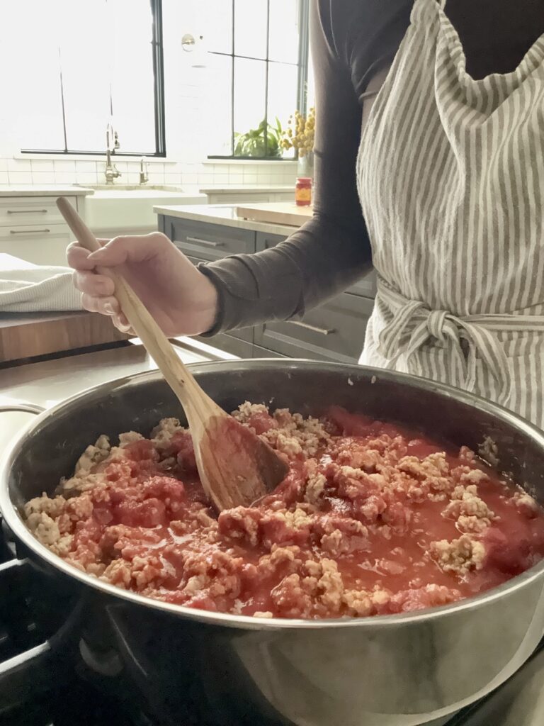 Home cook in a striped apron stirring ground chicken and Italian sausage with tomatoes in a stainless steel skillet for Easy Chicken Bolognese