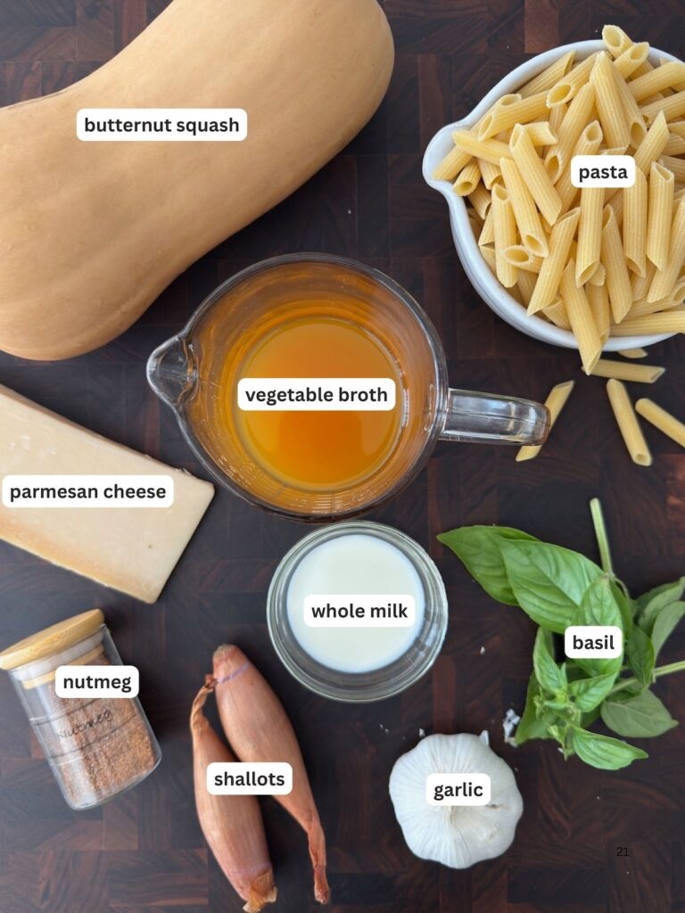 Ingredients for creamy butternut squash pasta sauce, including butternut squash, penne pasta, vegetable broth, Parmesan cheese, whole milk, fresh basil, shallots, garlic, and nutmeg on a dark background.