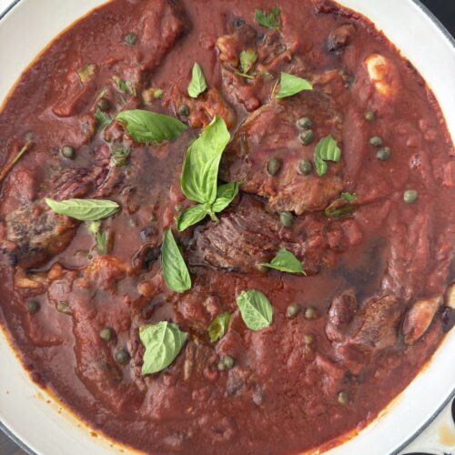 Steak Pizzaiola simmering in tomato sauce with fresh basil, capers, and olives in a white skillet