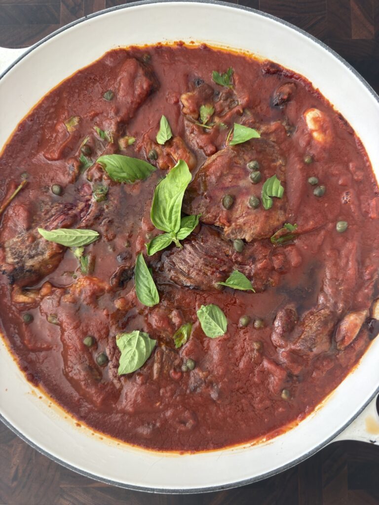 Steak Pizzaiola simmering in tomato sauce with fresh basil, capers, and olives in a white skillet