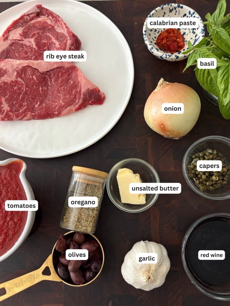 Overhead shot of labeled ingredients for Steak Pizzaiola, including ribeye steak, tomatoes, red wine, Calabrian chili paste, onion, garlic, olives, oregano, capers, basil, and butter