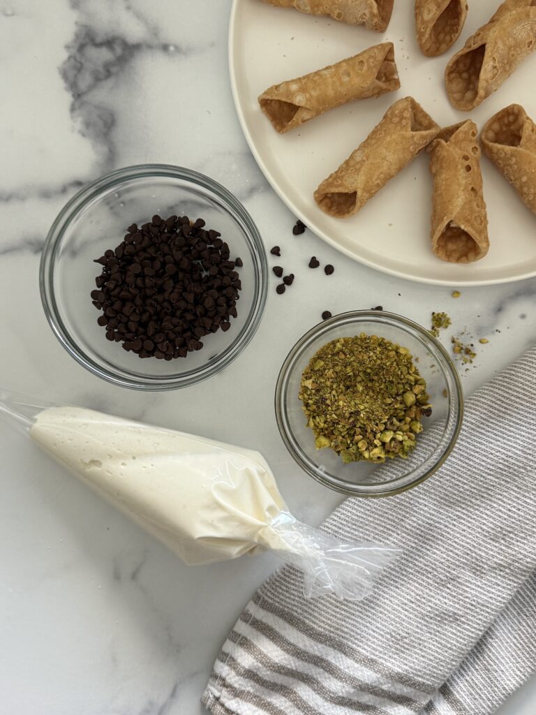 Cannoli shells on a plate with piping bag of ricotta filling, mini chocolate chips, and crushed pistachios ready for assembly.