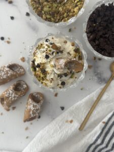 Bowls of cannoli cream topped with crushed pistachios and mini chocolate chips, served with spoons and cannoli shells for an Italian dessert.