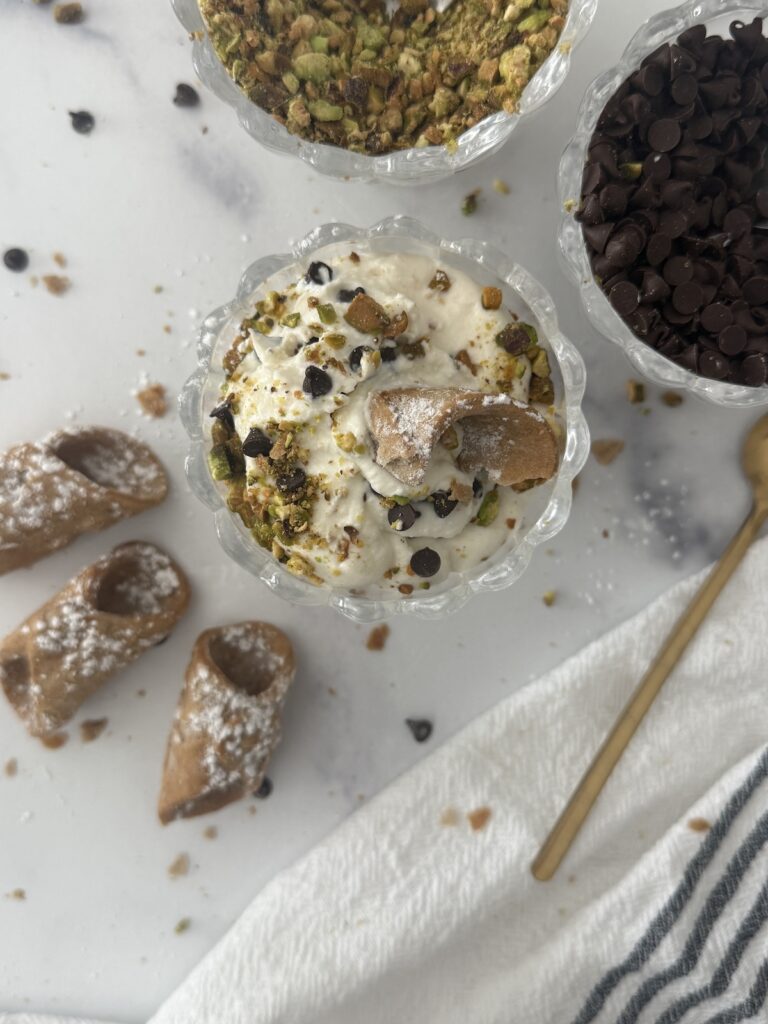 Bowls of cannoli cream topped with crushed pistachios and mini chocolate chips, served with spoons and cannoli shells for an Italian dessert.