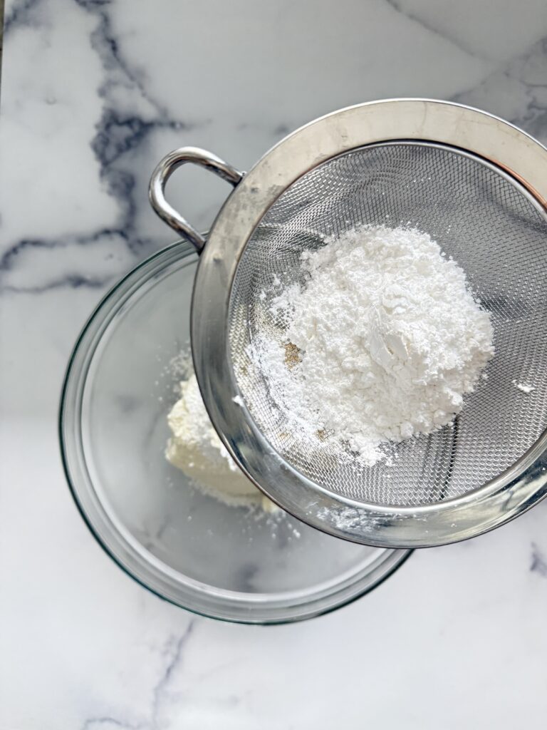 Sifting powdered sugar into a glass bowl with ricotta cheese to make creamy Italian cannoli filling