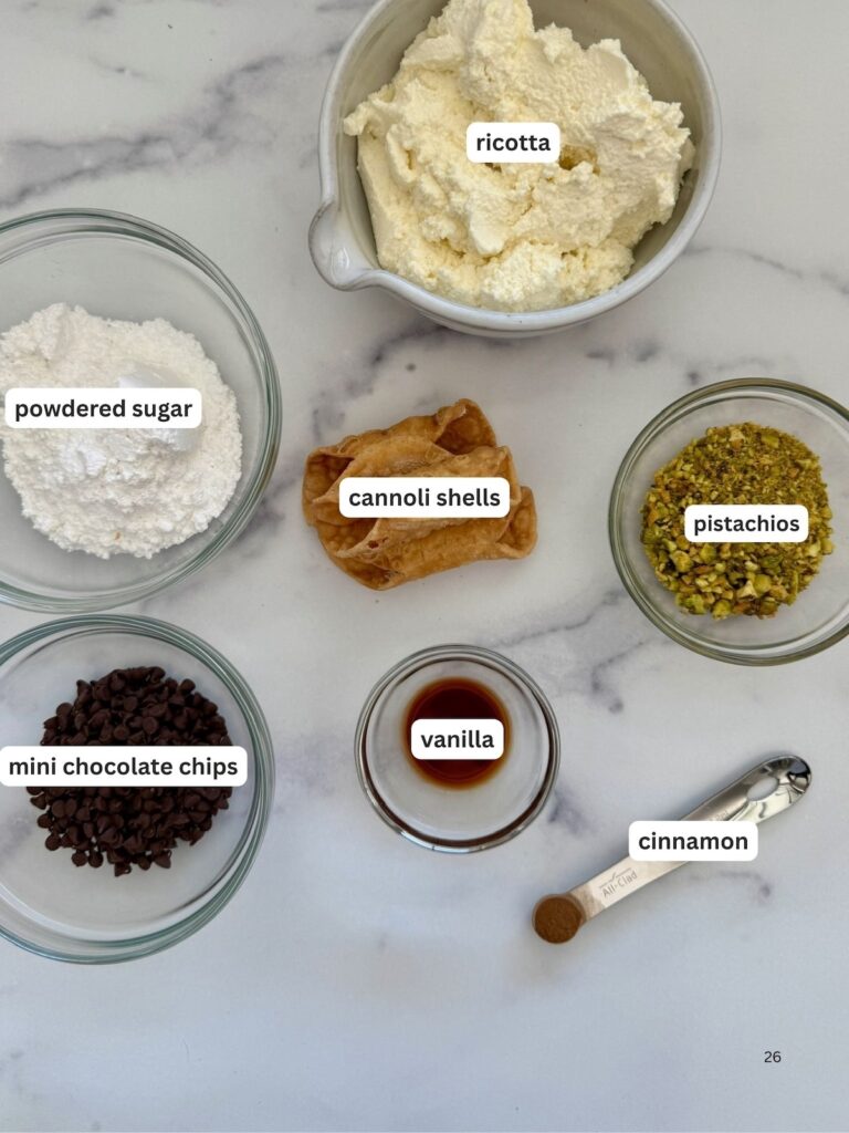 Cannoli ingredients on a marble background including ricotta cheese, powdered sugar, cannoli shells, chopped pistachios, mini chocolate chips, vanilla, and ground cinnamon for homemade Italian cannoli filling.