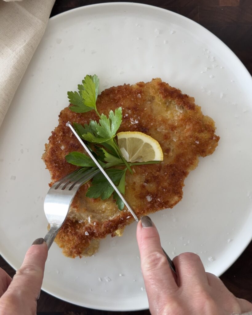 Crispy pork Milanese on a white plate topped with a lemon wedge and parsley, being cut with a fork and knife.
