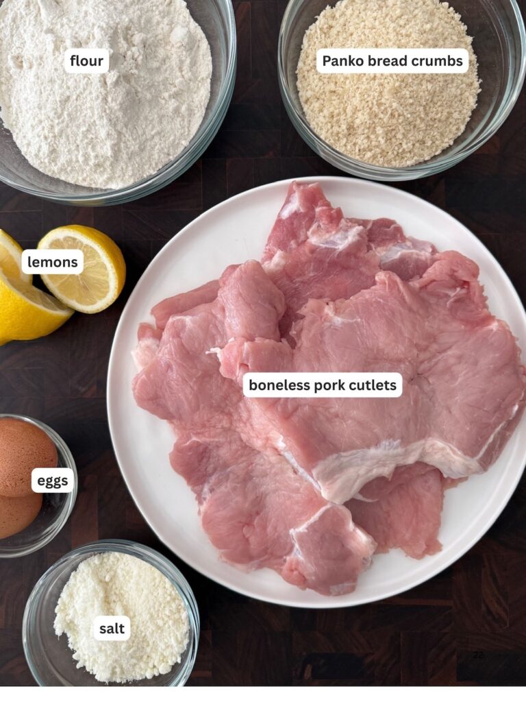 Overhead view of ingredients for pork Milanese including boneless pork cutlets, flour, Panko bread crumbs, eggs, lemons, and salt on a black background.