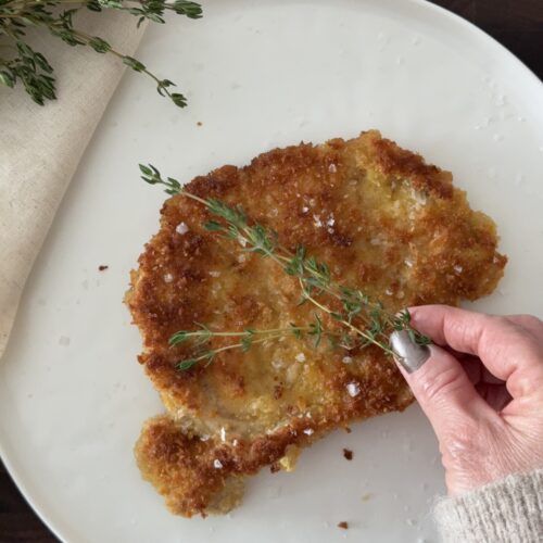 Crispy pork Milanese on a white plate being garnished with fresh thyme sprigs.