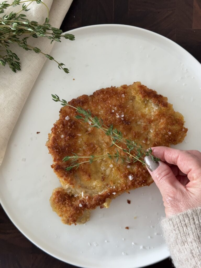 Crispy pork Milanese on a white plate being garnished with fresh thyme sprigs.
