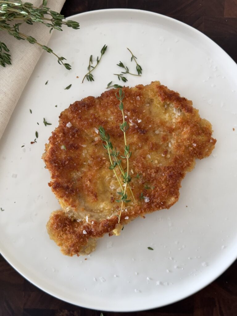 Crispy pork Milanese on a white plate being garnished with fresh thyme sprigs.