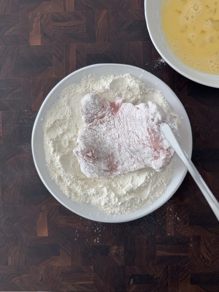 Boneless pork cutlet coated in flour during the dredging process for pork Milanese, with beaten eggs in a bowl nearby.