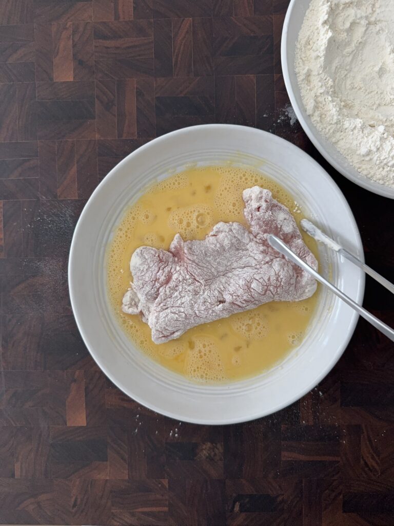 Boneless pork cutlet dipped in beaten eggs during the breading process for pork Milanese, with a bowl of flour nearby.