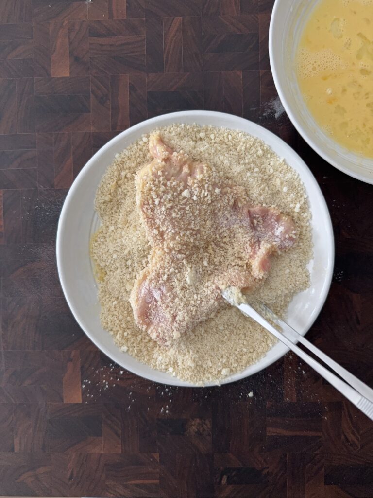 Boneless pork cutlet coated in Panko bread crumbs during the breading step for pork Milanese, with a bowl of beaten eggs nearby.