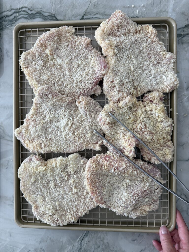 Overhead view of ingredients for pork Milanese including boneless pork cutlets, flour, Panko bread crumbs, eggs, lemons, and salt on a black background.