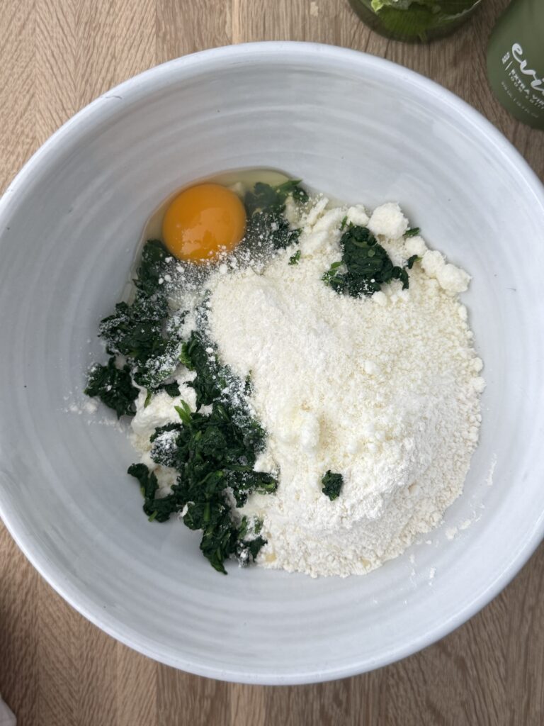 Ingredients for spinach and ricotta gnocchi in a white bowl, including egg, flour, ricotta, and cooked spinach.