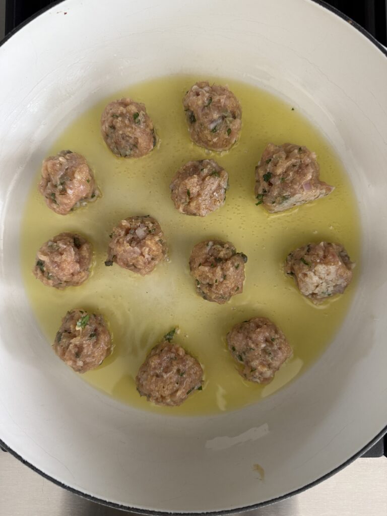 Chicken piccata meatballs searing in olive oil in a white skillet on the stovetop.