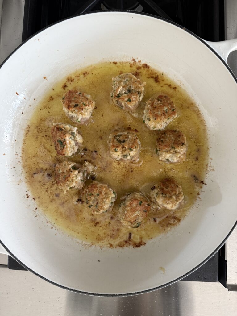 Chicken piccata meatballs simmering in lemon-caper butter sauce in a white skillet on the stovetop.