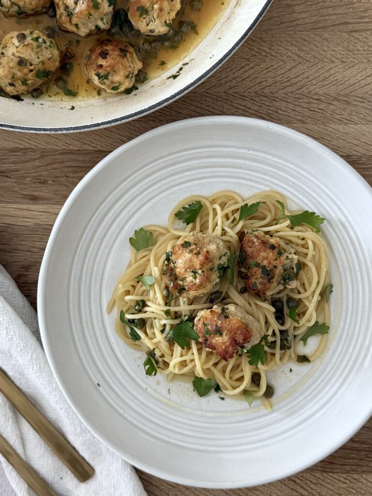 Chicken piccata meatballs with lemon-caper sauce over spaghetti, with skillet of meatballs in the background.