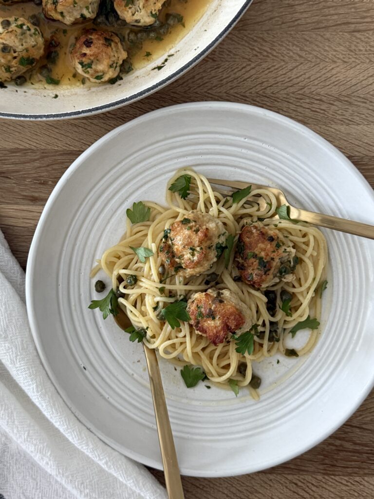 Chicken piccata meatballs over spaghetti with lemon-caper sauce and parsley, plated with the skillet in the background.
