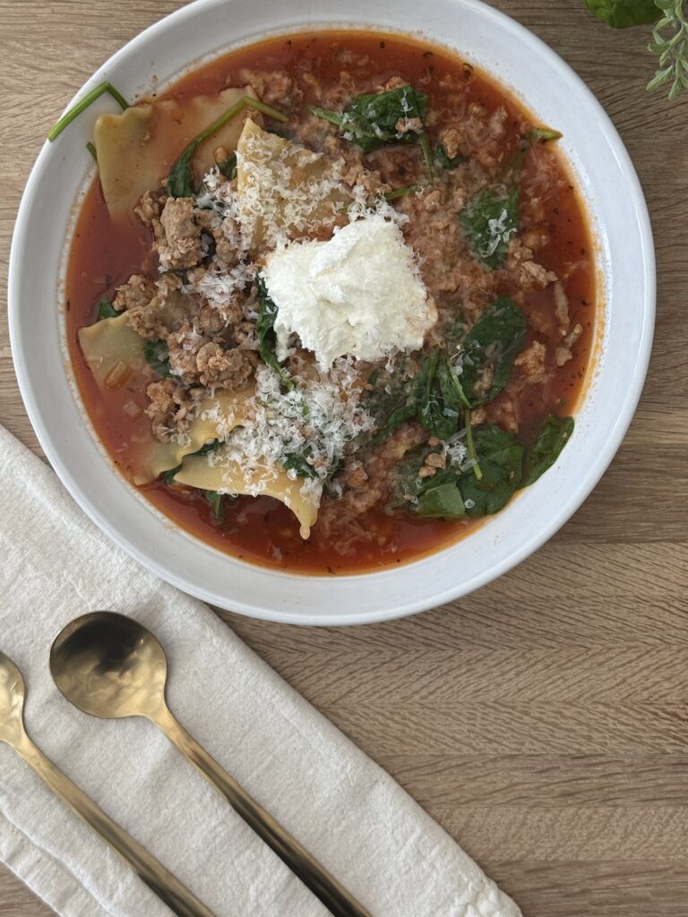 Lasagna soup in a white bowl garnished with ricotta and parmesan on a wooden board with two spoons and a cream napkin.