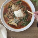 Lasagna soup in a white bowl garnished with ricotta and parmesan on a wooden board with a spoon in the bowl, ready to be eaten.