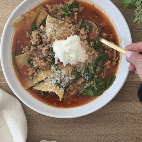 Lasagna soup in a white bowl garnished with ricotta and parmesan on a wooden board with a spoon in the bowl, ready to be eaten.
