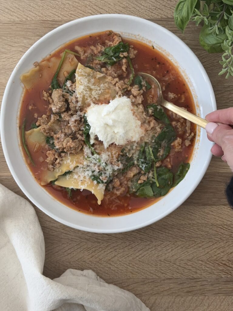 Lasagna soup in a white bowl garnished with ricotta and parmesan on a wooden board with a spoon in the bowl, ready to be eaten.