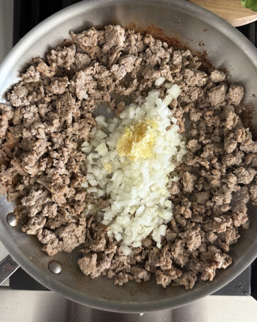 A photo showing the chopped onion and garlic with the browned meats in a large stainless skillet on the stove.