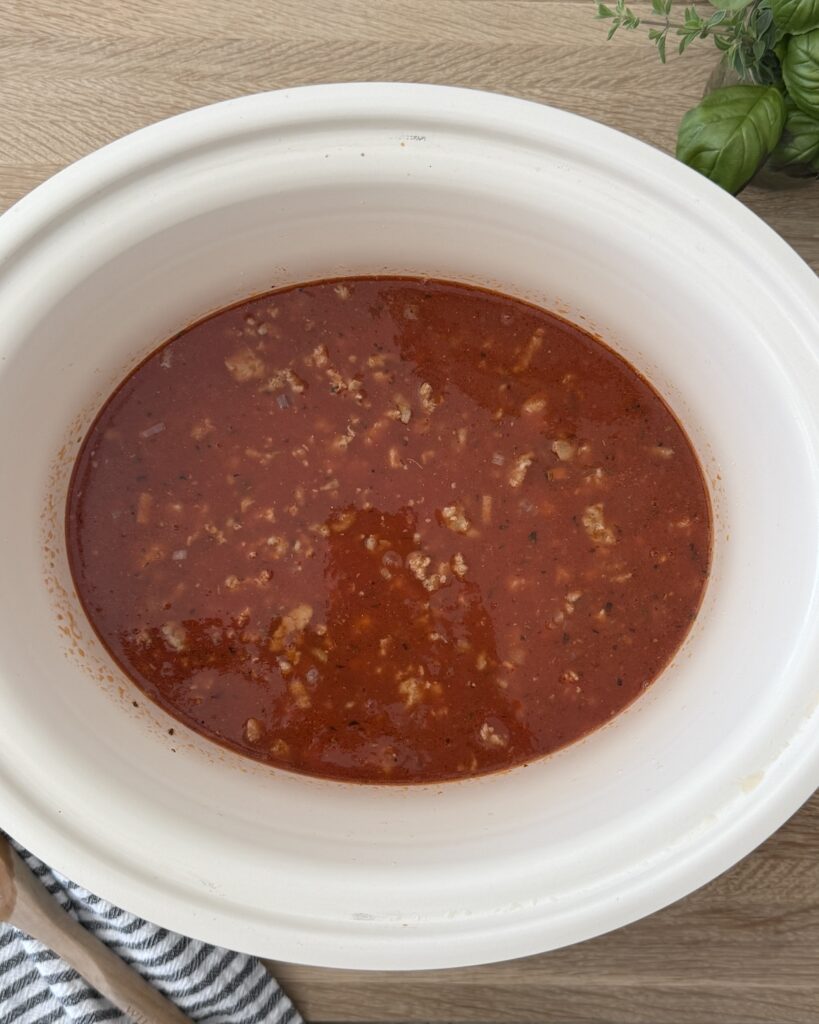A photo of the lasagna soup before noodles and spinach are added in the slow cooker on a wood cutting board.