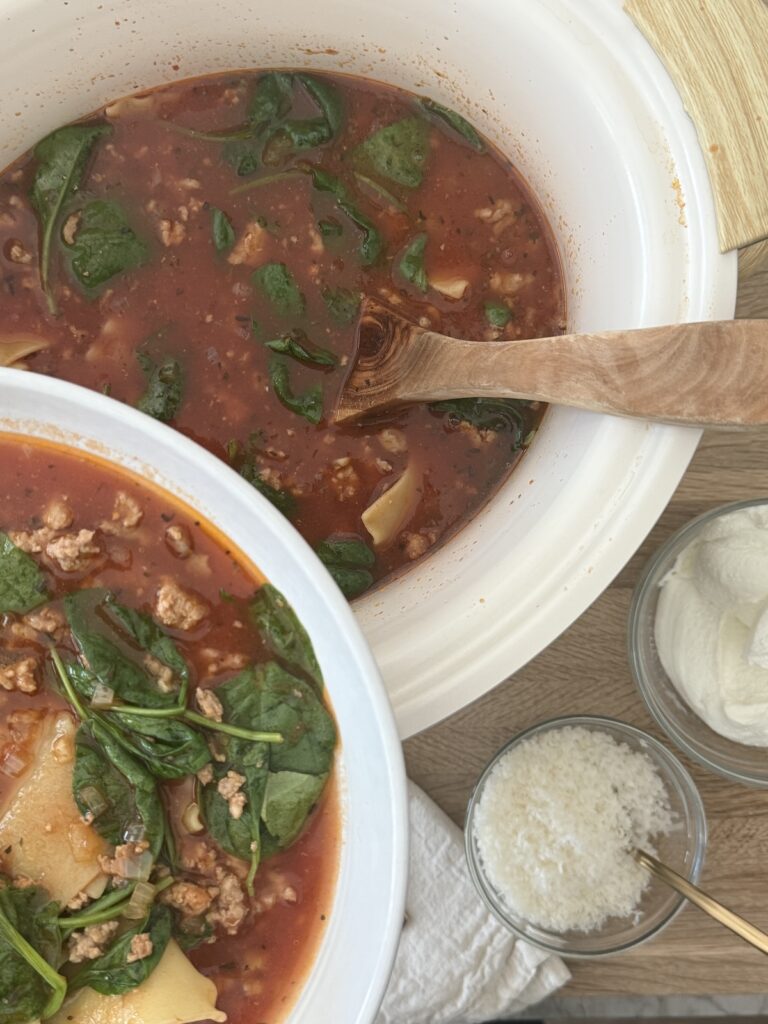 lasagna soup in the slow cooker, ready to be served with the grated parmesan and ricotta in bowls on a cutting board.