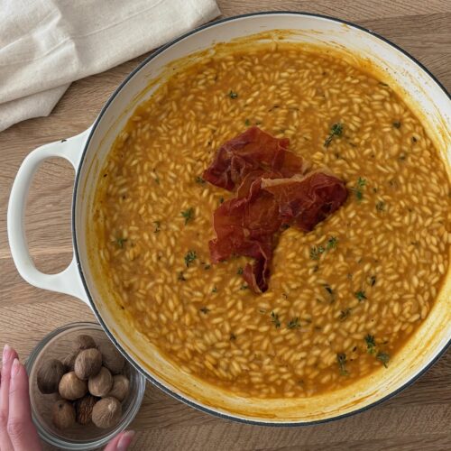 Creamy pumpkin risotto topped with crispy prosciutto and fresh nutmeg on a wooden board.