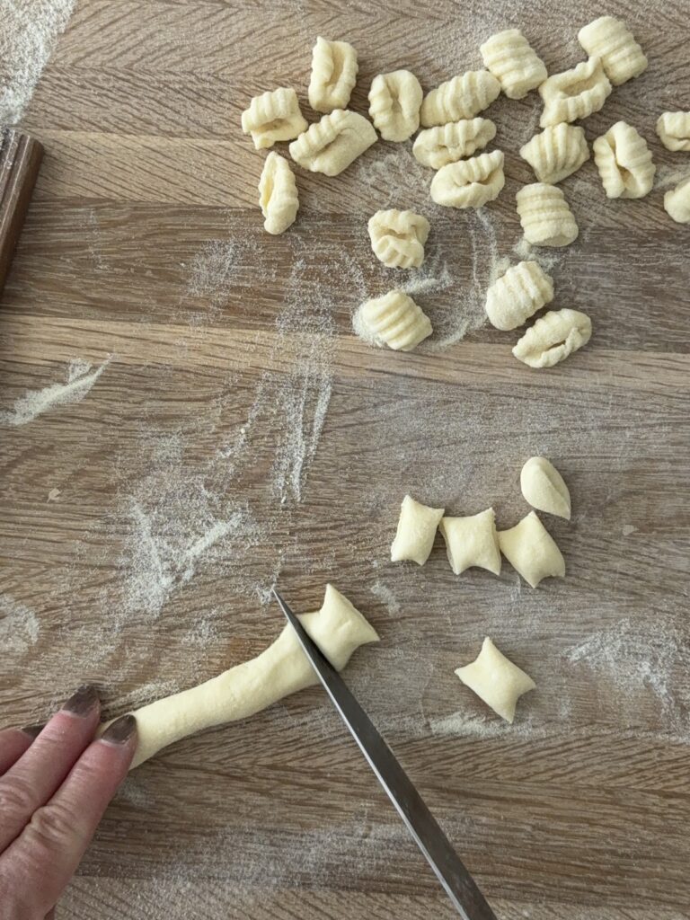 Hand shaping semolina pasta on a wooden gnocchi board beside freshly rolled pasta pieces on a floured surface.