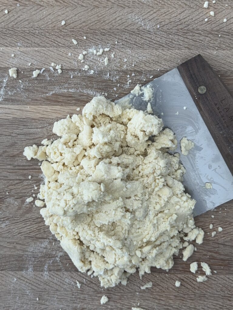 Shaggy-looking semolina pasta dough on a wooden surface with a bench scraper.