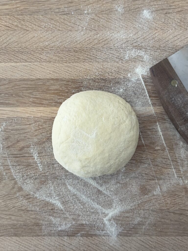 Ball of semolina pasta dough resting on a floured wooden surface with a bench scraper nearby.