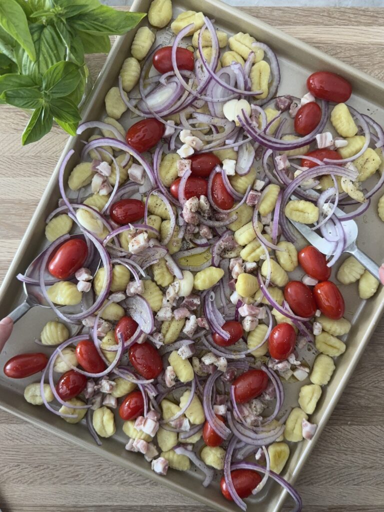 Uncooked sheet pan gnocchi with potato gnocchi, cherry tomatoes, sliced red onion, pancetta, garlic, and fresh basil arranged on a baking sheet — a simple Italian dinner ready to roast in the oven.