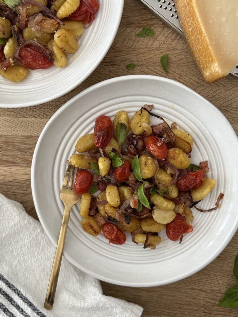 Sheet pan gnocchi with roasted cherry tomatoes, crispy potato gnocchi, red onion, pancetta, garlic, fresh basil, and Parmesan cheese served in a white bowl