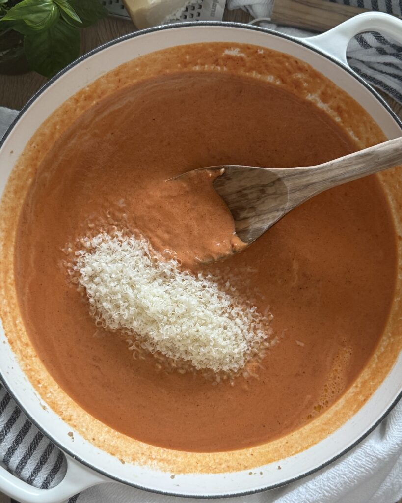 White Dutch oven filled with tomato cream sauce for baked rigatoni and Italian sausage, with grated Parmesan cheese being stirred in with a wooden spoon.