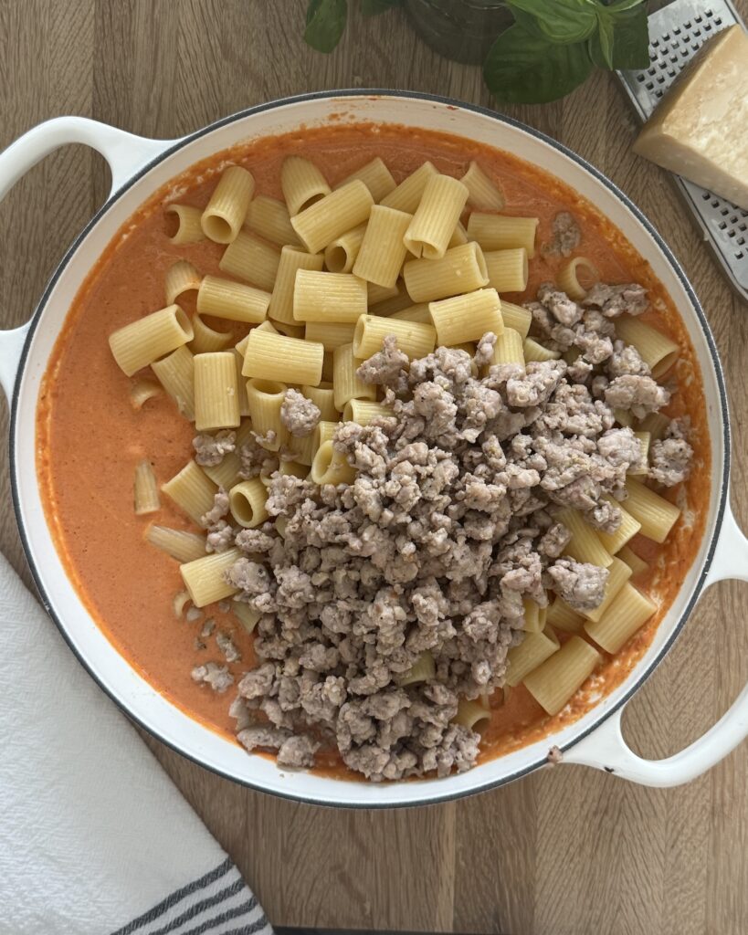 Dutch oven filled with tomato cream sauce, cooked rigatoni pasta, and browned Italian sausage, ready to be mixed for baked rigatoni and sausage.