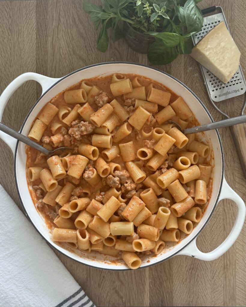 White Dutch oven filled with rigatoni pasta in tomato cream sauce, with a serving spoon, fresh basil, and a wedge of Parmesan on a wooden table.