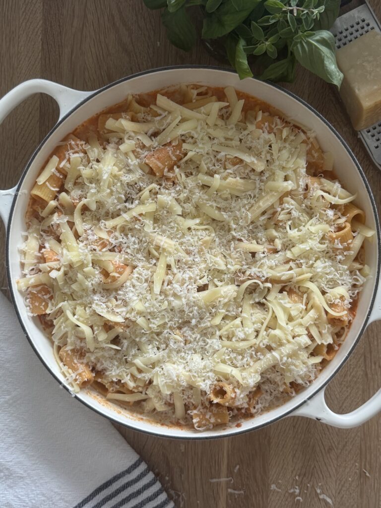 Unbaked baked rigatoni and Italian sausage in a white Dutch oven, topped with shredded mozzarella and grated Parmesan before baking.