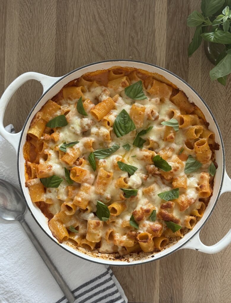 Overhead view of baked rigatoni and Italian sausage in a white Dutch oven, topped with melted mozzarella, tomato cream sauce, and fresh basil on a wooden table.