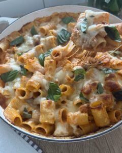 Overhead view of baked rigatoni and Italian sausage in a round pan, topped with melted mozzarella, tomato sauce, and fresh basil leaves.